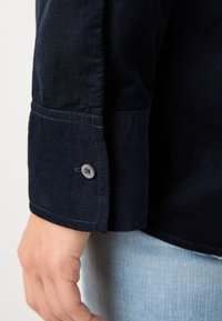 Gros plan sur le poignet d'une personne portant une chemise en velours côtelé bleu foncé avec un poignet boutonné et un jean denim bleu clair.