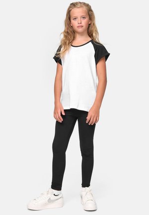 Niña joven con cabello largo y rubio, vestida con una camiseta de manga corta blanca y negra, leggings negros y zapatillas blancas, de pie contra un fondo liso.