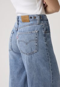 Άτομο που φοράει ανοιχτό μπλε τζιν Levi's με ορατές πίσω τσέπες και ετικέτα της μάρκας, στέκεται με σταυρωμένα τα χέρια φορώντας λευκό τοπ.