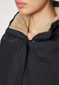 Veste noire avec un col en fourrure beige, dotée d'un zip à l'avant et de fermetures à pression. Logo Jack Wolfskin brodé sur la poitrine.