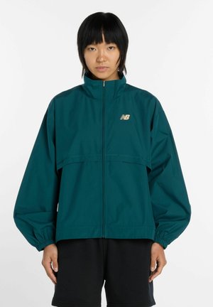 Persoon met kort zwart haar draagt een teal New Balance jas met rits en zwarte korte broek, staand tegen een effen witte achtergrond.