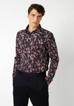 Mann trägt ein dunkles, langärmeliges, geblümtes Hemd und schwarze Hose, steht mit Händen in den Taschen vor einem schlichten weißen Hintergrund.
