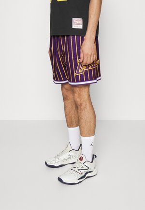 Mężczyzna w fioletowo-żółtych, pasiastych spodenkach do koszykówki Lakers, białych skarpetkach z logo Jordan i białych sneakersach New Balance, stoi w pomieszczeniu.