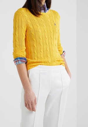 Vrouw die een geel kabelgebreide trui draagt over een opgerold geruit overhemd en witte broek met hoge taille, tegen een effen achtergrond.