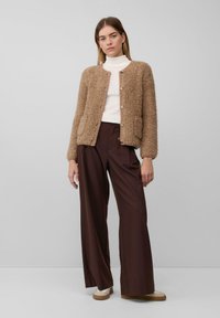 Vrouw in een beige gestructureerde button-up jas, witte coltrui, wijdbruine broek en beige schoenen, staand tegen een effen achtergrond.