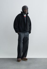 Schwarze Fleece-Reißverschlussjacke, dunkelgraue Cargohose, braune gemusterte Schuhe und ein brauner Helm. Gestylt mit einem schwarzen und roten Rollkragenpullover.