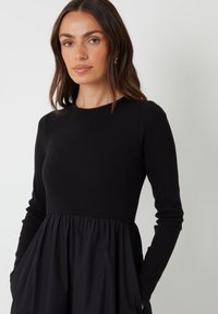 Abito nero con un corpetto a coste e una gonna ampia e arricciata. Le maniche lunghe e le tasche laterali aggiungono funzionalità al design.