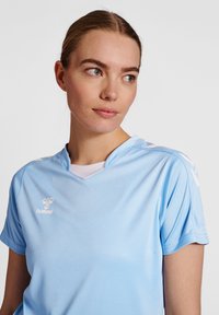 Camiseta atlética de color azul claro, fabricada con un tejido que absorbe la humedad. Presenta un escote en forma de V, mangas cortas y un logo blanco en el pecho y los hombros.