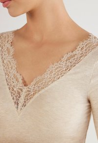 Beige topp med djup V-ringning och intrikata spetsdetaljer runt halslinningen. Tyget är slätt med en subtil textur.