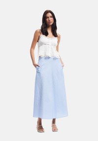 Top blanco con encaje y un corsé ajustado. Falda larga de cuadros azul con bolsillos. La modelo está de pie y lleva sandalias de tiras.
