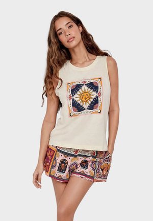 Femme aux longs cheveux ondulés, portant un débardeur avec un motif de soleil et un short à motifs représentant les signes du zodiaque Gémeaux et Scorpion.