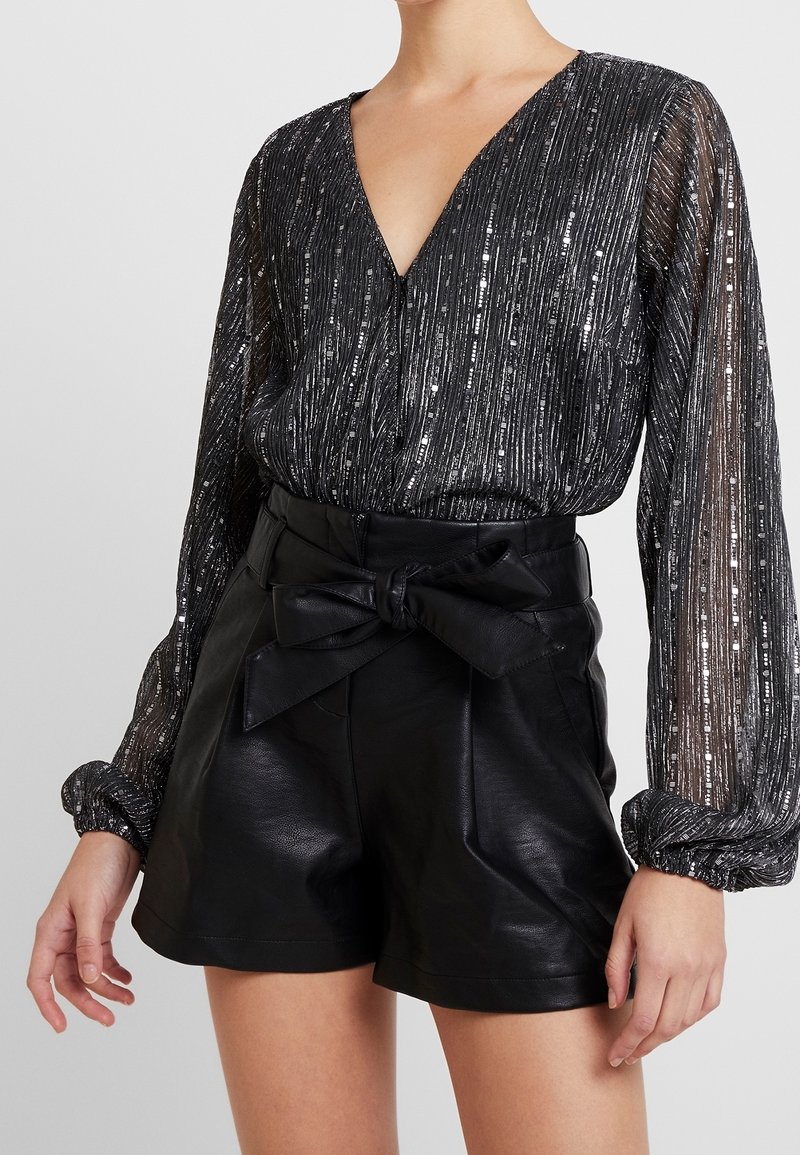 Blouse noire à manches longues en sequins avec col en V, associée à un short en cuir noir taille haute avec un grand nœud de ceinture.