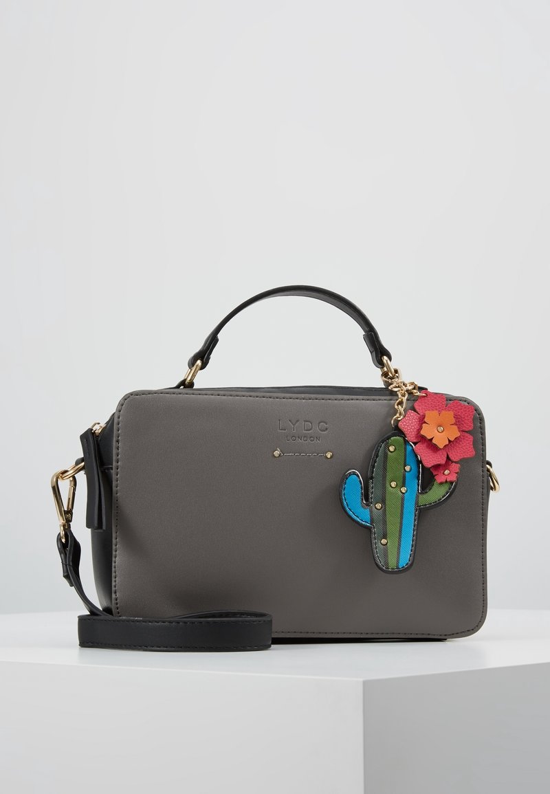 Sac à main rectangulaire gris avec poignée et sangle noires, garnitures dorées, logo de la marque, et porte-clés multicolore en forme de cactus avec une fleur rose attachée.