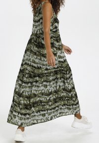 Maxi jurk met een groen en zwart abstract patroon, mouwloos ontwerp, gelaagde rok en lichte, vloeiende stof. Draagt het met witte sneakers.