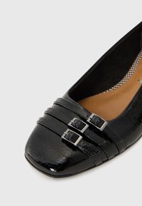 Chaussure plate en cuir verni noir avec un bout carré, présentant trois bandes horizontales et deux boucles en argent. Doublure intérieure beige.