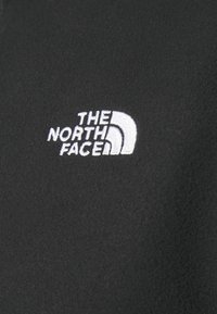 Schwarzer Fleece-Stoff mit einem weißen gestickten Logo von "THE NORTH FACE", das ein stilisiertes Halbkuppeldesign zeigt. Glatte Textur, minimalistisches Design.