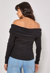 Schwarzes, langärmliges, schulterfreies Oberteil mit gerafften Stoffdetails am Kragen, kombiniert mit hellblauen, hochtaillierten Jeans.