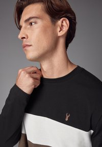 Košile s dlouhým rukávem s designem v černé, bílé a hnědé barvě, s texturovanou bílou částí a vyšívaným logem jelena na hrudi.