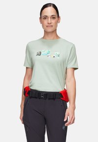 T-shirt verde chiaro a maniche corte con stampa grafica in più colori. Abbinato a pantaloni neri, con una cintura rossa in vita e tasche.