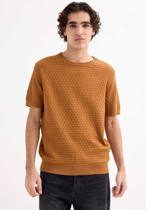 Suéter de manga corta y cuello redondo en mostaza con patrón de tejido texturizado. Puños y dobladillo de canalé, combinado con jeans oscuros.