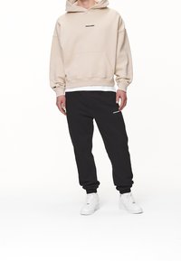 Beżowa bluza z kapturem i przednią kieszenią, czarne dresy z logo oraz białe sneakersy. Miękki materiał, luźny krój, minimalistyczny design.