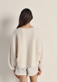 Kremas vilnos megztinis su nuleistais pečiais, tekstūruota medžiaga ir laisvu kirpimu, derinamas su pilkomis šortais.