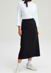Sort midi-nederdel med en glat tekstur og en elastisk talje, parret med en hvid langærmet top og hvide sneakers med grønne detaljer.
