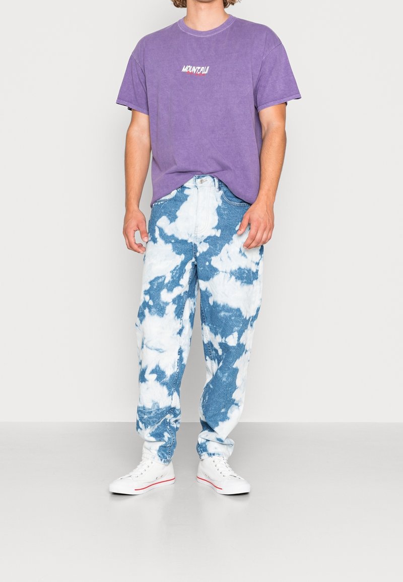 Lila bomulls-t-shirt med texten "MOUNT FUJI". Blå denimjeans med vitt molnmönster. Vita sneakers. En enkel, avslappnad outfit.