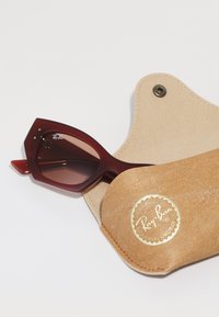 Ochelari de soare Ray-Ban cu ramă vinie, design unghiular și lentile maronii cu gradient, așezati într-un toc din piele maro deschis cu un logo auriu.