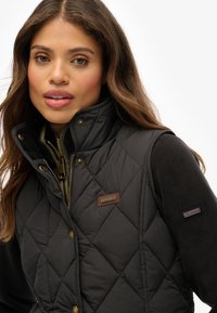 Zwarte gewatteerde puffer vest met hoge kraag, gouden hardware en een bruine merkembleem. Gelaagd over een geribbeld lichtkleurig vest.
