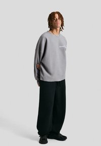 Šedý oversized sweatshirt s kulatým výstřihem, na kterém je malá bílá nášivka. Spárován s černými volnými tepláky a tmavými teniskami.