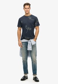 Camiseta azul marino con patrón floral, combinada con jeans de lavado claro y una camisa de rayas azul clara atada a la cintura; mocasines negros en los pies.