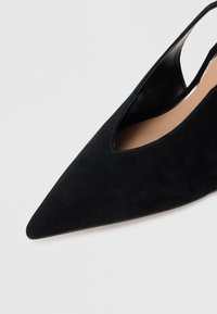 Czarny zamszowy sandał na pięcie z noskiem w szpic, z gładką jasnobrązową wkładką na tle jednolitego jasnego tła.