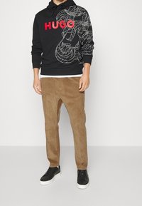 Černá mikina s kapucí s červeným logem "HUGO" a bílým liniovým designem; doplněná hnědými kordovými kalhotami a černými teniskami s bílou podrážkou.