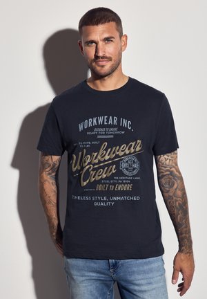 Hombre con tatuajes que lleva una camiseta impresa de trabajo color azul marino y jeans azules, de pie frente a una pared blanca con sombra.