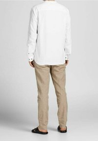 Vit långärmad skjorta med en avslappnad passform, kombinerad med beiga linnebyxor, sedd bakifrån. Svarta sandaler fulländar looken.