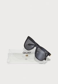Mustad musta värvi prillid tumedate läätsedega, mis puhkavad läbipaistvas karbis. Karbil on kuldne nupp ja logo "QUAY."