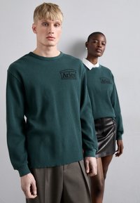 Green textured long-sleeve top with logo, paired with brown pleated pants on the left; model on the right in green knit top and leather skirt.