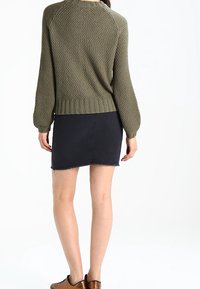 Pull en tricot vert olive avec un motif texturé, ourlet côtelé, associé à une jupe en denim noire avec un ourlet effiloché. Chaussures marron.