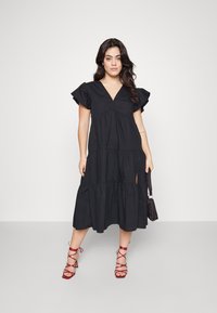 Black tiered midi dress with short ruffled sleeves, V-neckline, and side slit. Worn with red strappy heels and a small black handbag.