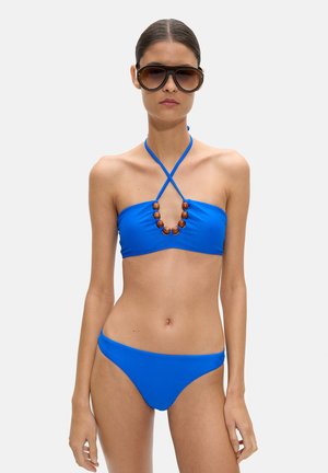 Kobieta w niebieskim bikini na szyję z drewnianymi koralikami i dużymi brązowymi okularami przeciwsłonecznymi, stojąca na tle jasnego, jednolitego tła.