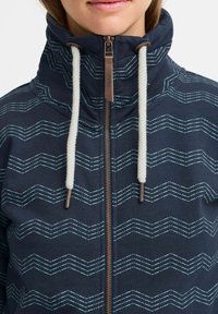 Marinblå jacka med dragkedja och hög krage, med blå vågmönster, vita snöre och en bronzfärgad dragkedja. Mjuk, texturerad tyg.