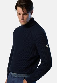 Marineblauer gerippter Rollkragenpullover mit langen Ärmeln, der ein Logo-Patch am linken Arm und ein tailliertes Design aufweist, kombiniert mit einem Gürtel.