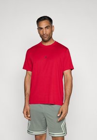 Κόκκινο βαμβακερό T-shirt με στρογγυλή λαιμόκοψη και κοντά μανίκια, με μικρό μαύρο λογότυπο στο στήθος. Φοριέται με γκρι αθλητικά σορτς.