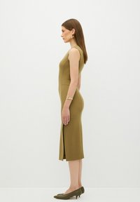 Olijfgroene, getailleerde mouwloze jurk met een zijsplit, een gladde textuur en een bescheiden zoom, gecombineerd met groene hakken en gouden oorbellen.