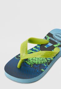 Green rubber flip-flop with textured straps, a blue and black sole featuring a cartoon design, and a light blue base. Accessible design.