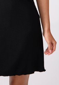 Czarna sukienka o gładkiej fakturze, z falbaniastym dołem i dopasowanym krojem. Ręka spoczywa w pobliżu, z neutralnym lakierem do paznokci.