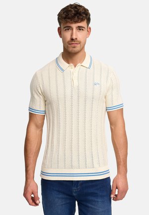 Jonge man die een crèmekleurige gebreide polo draagt met blauwe strepen op de kraag, mouwen en zoom, gecombineerd met een blauwe spijkerbroek, gezicht naar voren gekeerd tegen een effen achtergrond.