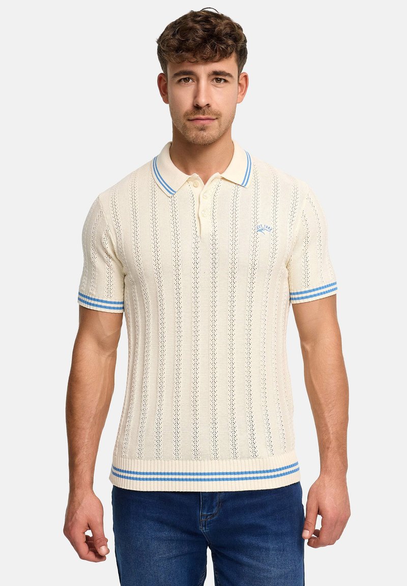 Jeune homme portant un polo en maille écru avec des rayures bleues sur le col, les manches et l'ourlet, associé à un jean bleu, faisant face à l'avant sur un fond uni.