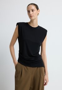 Černý top bez rukávů s kulatým výstřihem, zdobený splývavou přední částí a hladkým povrchem, doplněný o volné hnědé kalhoty.